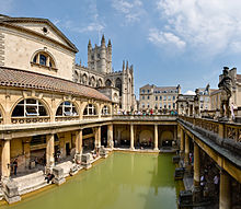 Photo des Bains montrant une zone rectangulaire de l'eau verd??tre entour??e de b??timents en pierre jaune avec des piliers. Au fond, la tour de l'abbaye.