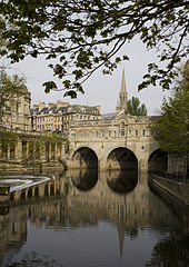 Jaune Gris pont / en pierre avec trois arches sur l'eau qui refl??te le pont et le clocher de l'??glise derri??re. Un barrage est sur la gauche avec d'autres b??timents en pierre jaune derri??re.