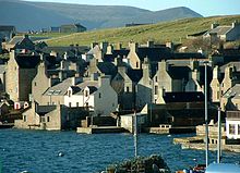 Les maisons de pierre se pressent autour d'un rivage, le pignon extr??mit??s face ?? l'eau, avec des collines vertes au-del??.