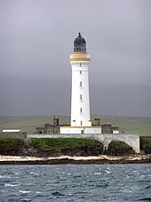 Un grand phare blanc avec une bande brune autour du parapet et la lanterne de couleur sombre se asseoir sur une c??te rocheuse. Un mur blanc obscurcit l'??tage inf??rieur de b??timents de pierre grise r??unis autour de sa base.