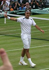 Un homme aux cheveux noirs est saluant la foule avec sa raquette de tennis dans sa main droite, et il porte tous les v??tements blancs