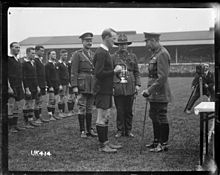 Une photo en noir et blanc d'un terrain de rugby dans lequel trois hommes en uniforme militaire, dont l'un est le roi George, pr??sentent un troph??e d'argent ?? un joueur de rugby habill?? en kit noir. Derri??re en ligne sont le reste de l'??quipe.