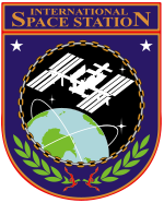 Une silhouette de l'ISS en orbite montr?? dessus de la Terre. Cette image est suspendu dans un bouclier orange et violet, avec de la Station spatiale internationale ??les mots ci-dessus l'image, et de feuilles de laurier-dessous.