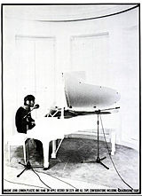 Une photo noir et blanc de Lennon assis ?? une salle blanche piano ?? queue. Il porte des ??couteurs et une chemise sombre.