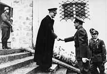 Photo noir et blanc, prise le 9 juin 1941 à Berchtesgaden, en Allemagne. Au centre de la photo, Adolf Hitler, portant un long manteau noir et une casquette militaire, sert la main d’Ante Pavelić (à droite), dirigeant de l’État indépendant de Croatie. Les deux hommes sont de profil, debout, sur les marches de l’escalier menant à l’entrée de la résidence secondaire de Hitler. Un soldat en arme se tient debout, en haut de l’escalier (en haut à gauche de la photo). Au second plan, deux officiers allemands (en bas à droite de la photo) assistent à la scène. L’arrière-plan de la photo est constitué d’un mur blanc percé d’une fenêtre grillagée (au-dessus de la tête du dirigeant croate).