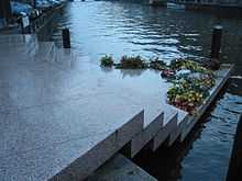 Photo en couleurs (teinte bleutée dominante), prise à Amsterdam en novembre 2003, et montrant le « Homomonument » : un monument dédié aux victimes homosexuelles du nazisme. La photo en montre seulement une partie : un assemblage de cinq dalles en granit rose qui forment un escalier triangulaire permettant d’accéder, du trottoir, au bord d’un canal (de gauche à droite sur la photo). La dernière marche, une plate-forme triangulaire, qui semble flotter sur l’eau du canal, est en partie recouverte de gerbes de fleurs. L’arrière-plan de la photo est constitué par l’étendue d’eau du canal.