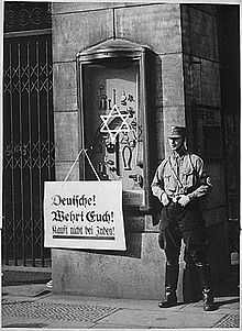 Photo noir et blanc du 1er avril 1993. Sur une vitrine, encastrée dans un mur et exposant des outils de bricolage, est accrochée une pancarte, sur laquelle on peut lire, en noir sur fond blanc : « Deutsche! Whert euch! Kauft nich bei juden! ». Un milicien, en uniforme de la SA, se tient debout à côté de la vitrine (à droite sur la photo), mains dans son ceinturon et jambes écartées. Une étoile de David blanche est peinte au centre de la vitre de la vitrine.