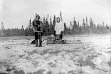 Deux hommes habillés de vêtements de fourrure posent devant un traineau. Le sol est neigeux. À lʼarrière plan, une ligne dʼépinettes noires.