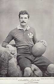 Photo portrait d'Arthur Gould vêtu du maillot gallois. Il est assis et retient de la main un ballon de rugby posé sur sa cuisse gauche.