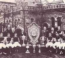 Photo en noir et blanc d'une équipe de football posant autour de son blason et de ses trophées au centre.