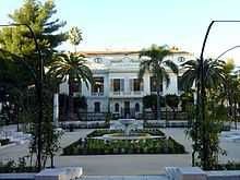 Vue d'une place avec fontaine et au fond un bâtiment de type colonial.