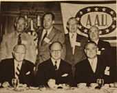 Des hommes assis à une table avec, en arrière-plan, des hommes debout et un drapeau « A.A.U. »