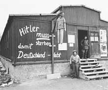 Photo noir et blanc de l’entrée d’une baraque en bois dans le camp de concentration de Buchenwald. Au centre de la photo, près du mur de la baraque, se dresse une potence à laquelle pend un pantin à l’effigie d’Adolf Hitler. Sur le mur de la baraque est peinte l’inscription en allemand : « Hitler muss sterben damit Deutschland lebt ». À droite, un homme, en tenue sombre, se tient debout dans l’encadrement de la porte, au sommet de l’escalier en bois qui mène à l’entrée. Un soldat américain pose debout à côté de l’escalier.