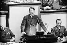 Photo noir et blanc prise le 4 mai 1941, à l’intérieur du Reichstag. Au centre de la photo, Adolf Hitler, debout, mains appuyées de part et d’autre d’un pupitre noir supportant 4 micros, prononce un discours. Il porte une cravate noire, une chemise blanche et une veste croisée sombre. Deux officiers sont assis à ses côtés. L’arrière-plan est constitué de trois panneaux de couleur claire.