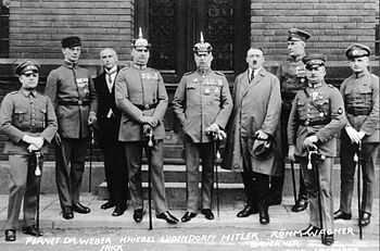 Photo noir et blanc de neuf hommes debout, dans une rue, devant la porte d'entrée fermée d'un bâtiment. Sept hommes sont en tenue militaire. Il s'agit d'Adolf Hitler et des autres personnalités inculpées lors du son procès en 1924.