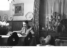 Photo noir et blanc montrant Adolf Hitler, assis à une table, dans une pièce de sa résidence secondaire de l’Obersalzberg. L’homme, en costume clair, de face, est en train de lire des documents. L’arrière-plan est divisé par un rideau aux motifs fleuris : à droite, une fenêtre fermée au bord chargé de pots de plantes et, à gauche, un mur blanc supportant un tableau et un meuble en bois d’une couleur foncée sur lequel sont posées, sur la tranche, trois assiettes.