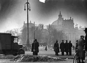 Photo noir et blanc sur laquelle le Reichstag, partiellement caché par une rangée d'arbres, est visible en arrière-plan, le 28 février 1933, lendemain de son incendie. Quelques badauds, en habits sombres, se tiennent debout au premier plan.