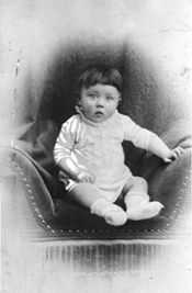 Photo noir et blanc d’Adolf Hitler prise peu après sa naissance. Sur un fond blanc, dans un médaillon au fond sombre, au centre de la photo, un bébé, en layette blanche, est assis sur un siège en velours foncé. Les yeux grands ouverts, il fixe l’appareil photo avec curiosité.