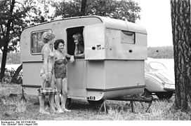 Photo noir et blanc de 1958 ; trois femmes discutent sur le pas d'une petite caravane blanche installée sous des arbres, non loin d'un lac.