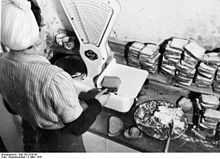 Photo d'une femme vue de dos pesant deux tranches de pain sur une balance placée devant elle.  À droite de la balance, une assiette de beurre sur laquelle repose le couteau à tartiner et des piles de tartines fourrées.