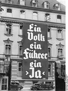 Photo noir et blanc, prise à Berlin en novembre 1933, d'une affiche électorale du NSDAP déployée sur les trois premiers étages de la façade d'un bâtiment de cinq étages (bureau du Parti nazi). On peut lire, en grosses lettres, le slogan en allemand : « Ein Volk, ein Führer, ein Ja ».