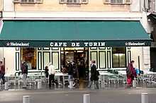 Vue de face d'un café avec son store vert tiré sur lequel est écrit en blanc « Café de Turin » en lettres majuscules.