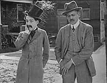 Capture d'écran montrant deux hommes en costume dans une rue. Chaplin porte un haut-de-forme et une moustache.