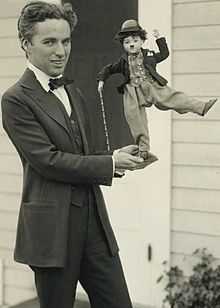 Photographie de Charlie Chaplin debout et en costume tenant une poupée représentant Charlot