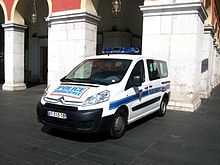 Vue d'une fourgonnette blanche de police municipale des années 2000 garée au pied d'arcades donnant sur une place.