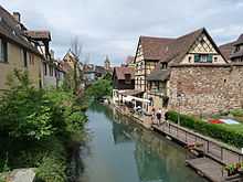 Maisons à colombages au bord d'une rivière. À droite, un quai ; à gauche des arbres ; en arrière plan, on aperçoit un clocher.