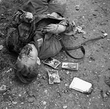 Photo noir et blanc prise le 23 décembre 1943 à Ortona, en Italie. Dans la partie gauche de la photo, le cadavre d’un soldat allemand gît (tête vers le bas de la photo) sur un sol de terre pierreuse. Trois photos de soldats sont visibles près de son épaule droite.