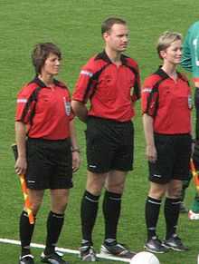 Photo de la présentation des trois arbitres en tenue (dont deux femmes) avec un ballon posé au sol.