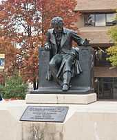 Statue d'un homme assis sur une chaise posée sur un socle de pierre au milieu d'un parc