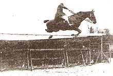 Photo en noir et blanc d'un homme franchissant un obstacle sombre surmonté d'une barre blanche; la position du cavalier est notable car il est très assis dans sa selle, les bras tendus et les rênes longues pour permettre au cheval de sauter.