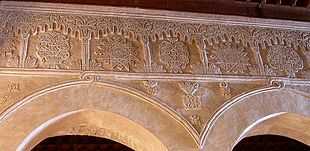Photographie de la frise, à décor en plâtre sculpté, qui surmonte les arcs moulurés de la nef centrale de la salle de prière. Cette frise, rythmée d’arcs à lambrequins, est composée de médaillons ornés de motifs géométriques et floraux répétitifs.