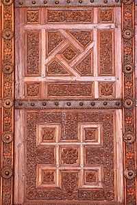 Photographie d’un vantail en bois sculpté de la grande porte de la salle de prière. Des bandes de tôle, ornées de clous à tête ronde, séparent les panneaux ouvragés de chaque vantail.