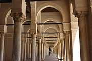 Vue intérieure du portique occidental, montrant une succession d’arcades. Celles-ci sont soutenues par des colonnes à chapiteaux de styles différents.