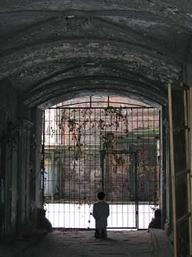 Photo en couleurs prise dans le hall d’un des bâtiments de l’ancien Ghetto de Varsovie en 2013. Sous un porche de pierre peu éclairé, on distingue la silhouette d’un homme agenouillé, de dos, devant une large et haute porte grillagée donnant sur une ruelle au sol très clair. Il s’agit d’une statue en cire, de l’artiste italien Maurizio Cattelan, représentant Adolf Hitler en costume gris, priant agenouillé.