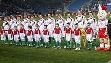 Sur un terrain de football, onze joueurs chantent leur hymne national, la main sur le cœur devant onze enfants.