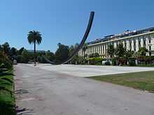 Vue d'une allée de parc dégagée avec palmier et sculpture contemporaine et une façade blanche imposante en fond.