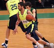 Navarro, en jaune, en mouvement et en dribble. Un défenseur, lgèrement en arrière, essaye de le retenir. Au fond de l'écran, un autre joueur en jaune, de dos.