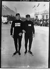 Photographie en noir et blanc de deux patineurs à l'arrêt au centre de la glace, se tenant pars le bras, portant une tenue sombre et un bonnet ainsi que le drapeau finlandais sur la poitrine.