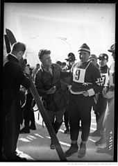 Photographie en noir et blanc d'un homme portant une tenue sportive avec le dossard numéro 9 et un bonnet, entouré d'autres hommes dont un face à lui tenant une paire de ski.