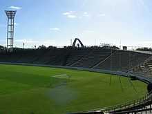 Stade de football avec une pelouse verte sans aucun joueur, sous un ciel bleu.