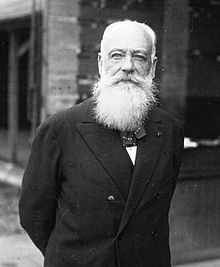 Portrait en buste d'un homme portant un costume complet et une barbe blanche, les bras croisés dans le dos.