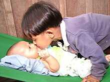 Sur la droite un jeune garçon d'apparence asiatique, les cheveux coupés en bol, se penche au-dessus d'un bébé allongé sur le dos à gauche de l'image. Le garçon et le bébé se touchent par le nez. Le bébé regarde le garçon avec une expression d'intense intérêt.