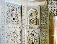 Photographie montrant quelques-uns des panneaux en marbre blanc, sculptés et ajourés, de la paroi du mihrab. Entre les panneaux est visible une partie de l’inscription coranique en caractères coufiques.