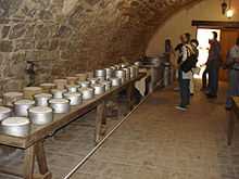 La photographie couleur représente une cave voutée en pierre. Une longue table en bois sur des tréteaux porte des fromages frais dans des moules métalliques et en terre cuite. Au fond de la salle, des visiteurs examinent des ustensiles en aluminium : bidons de lait et marmites de grand volume.