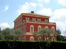 Vue d'un bâtiment en rouge avec façade en trompe-l'œil sous un ciel bleu avec deux arbres et des haies en premier plan.