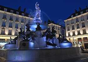 La fontaine, de nuit, éclairée en bleu sur fond d'immeubles éclairés en blanc.